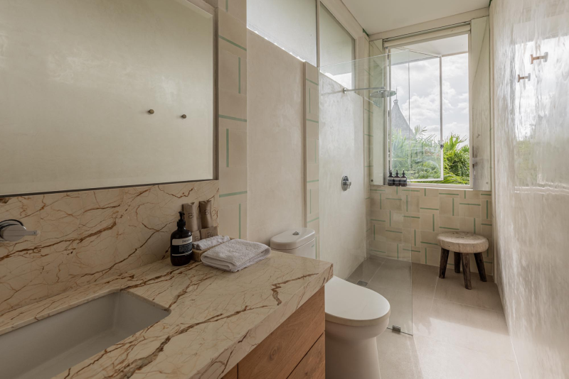 Bathroom with marble countertop, white sink, toiletries, toilet, glass shower, stool, large window with greenery outside, light-colored tiles, and sheer curtains.