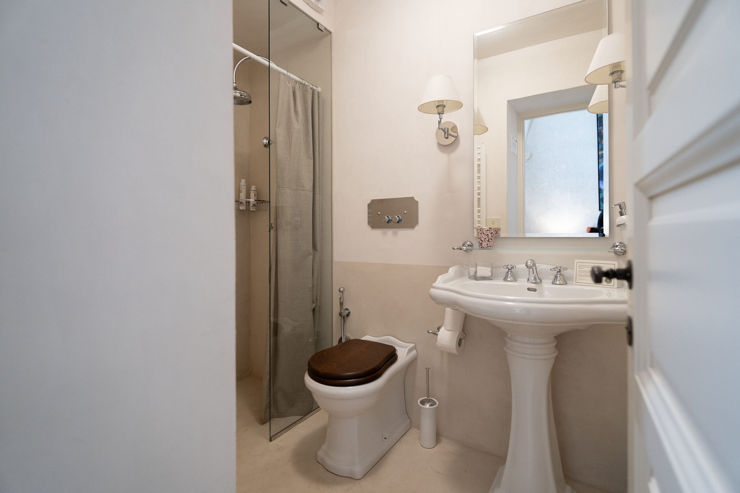 A bathroom with a shower area enclosed by a glass door, a toilet with a wooden seat, a pedestal sink with a mirror, and a window with a frosted glass pane.