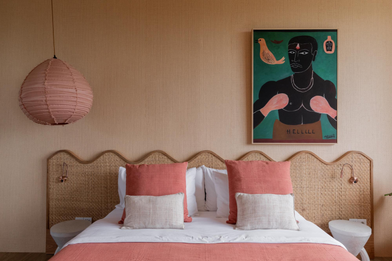 A cozy bedroom with a rattan headboard, white bedding, and pink throw pillows. A pink paper lantern hangs from the ceiling, and artwork depicting a figure with boxing gloves is on the wall.