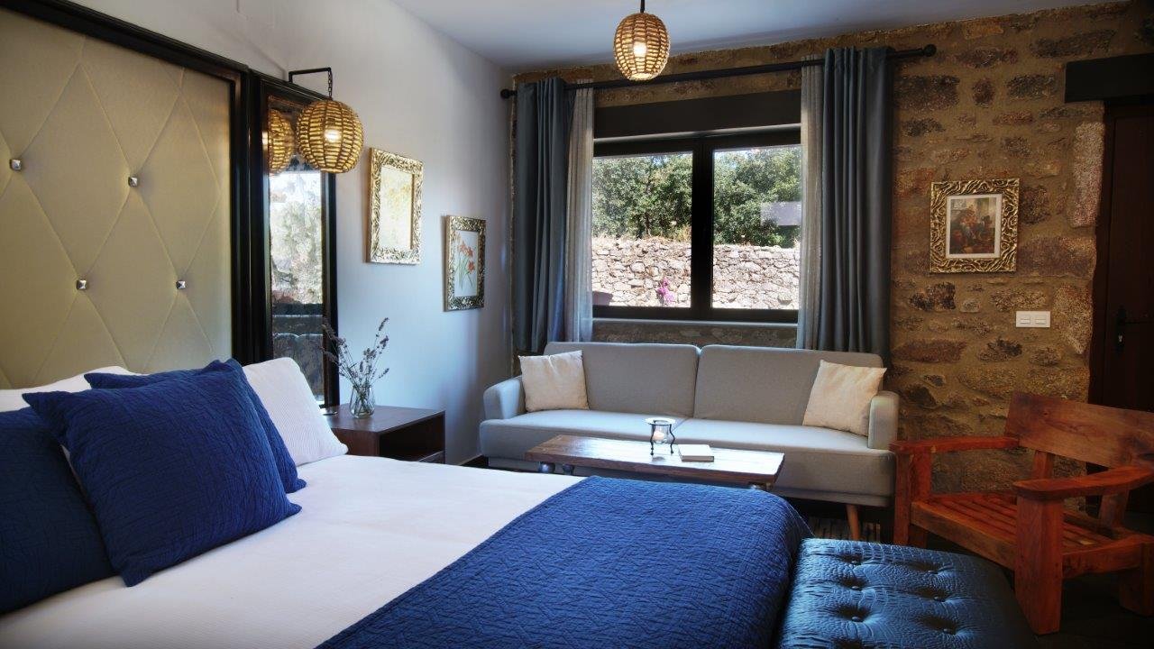 Hotel bedroom with a bed, a white sofa, wooden chair, framed art, window with curtains, and decorative lighting