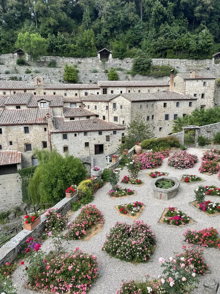 Stone buildings with tiled roofs, overlooking a flower garden with pink and red roses, a small fountain, and a green hillside in the background.