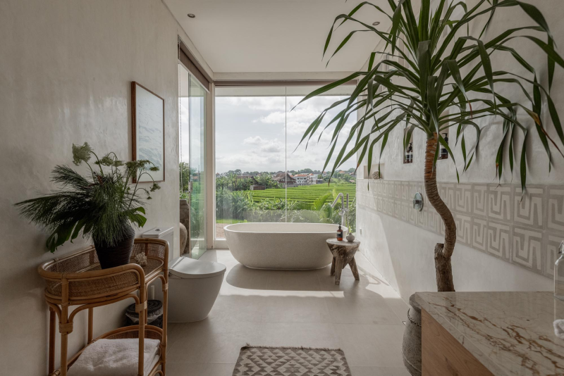 Modern bathroom with a freestanding bathtub facing large windows with a view of a lush green landscape and distant houses, decorated with indoor plants and natural materials.