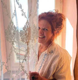 A woman with curly hair wearing glasses, earrings, and a striped shirt, smiling in a well-lit indoor setting.