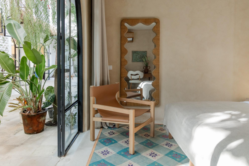 A cozy bedroom corner with a wooden armchair, a tall decorative mirror, a bed with white linens, a colorful patterned rug, and large glass doors opening to outdoor greenery.