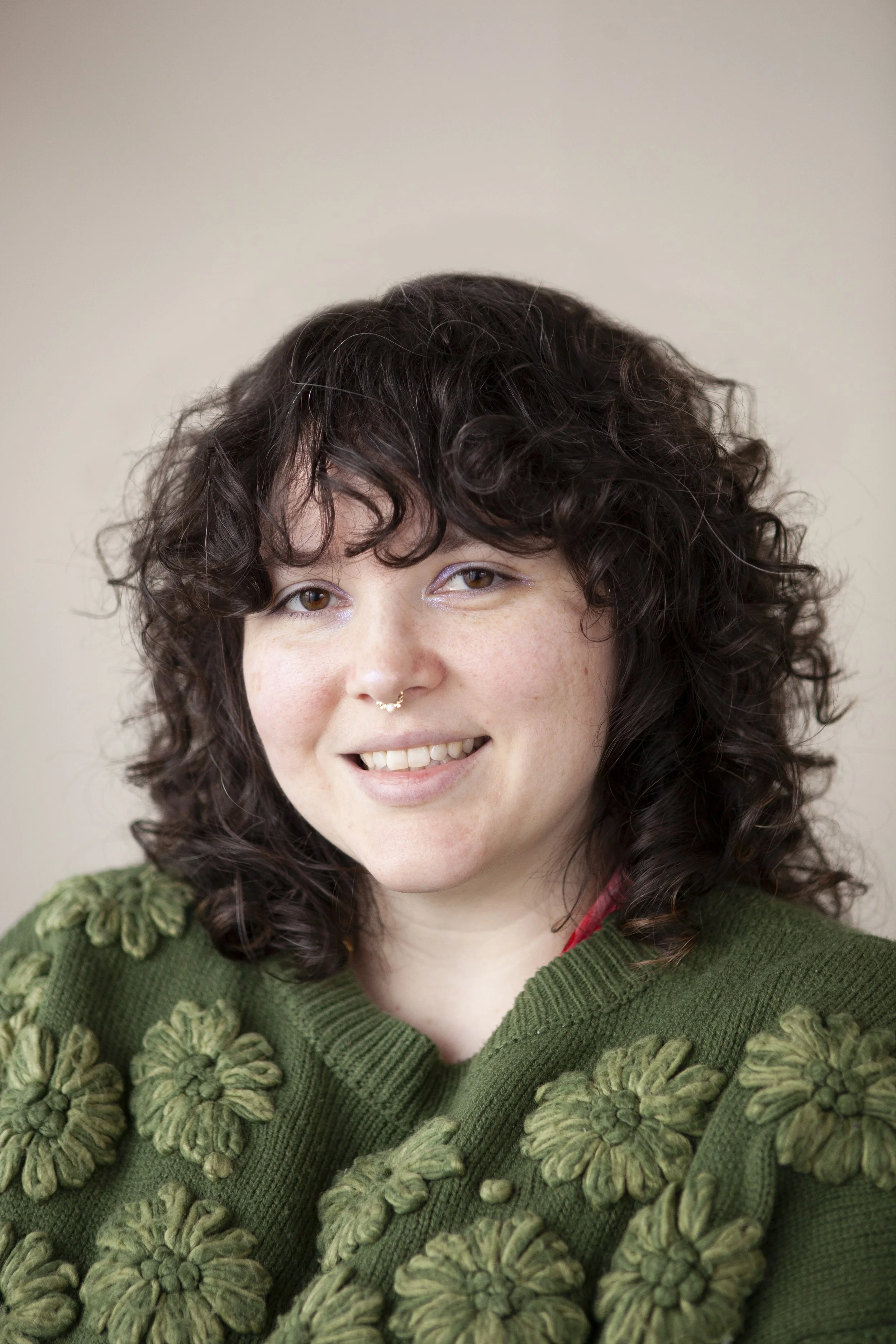 A woman with dark curly hair, wearing a green sweater with floral embroidery, smiling with a neutral background.