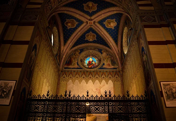 Interior view of a church or cathedral with ornate religious paintings, stained glass window depicting a religious figure, decorative ceiling with gold accents, and a wrought iron gate in the foreground.