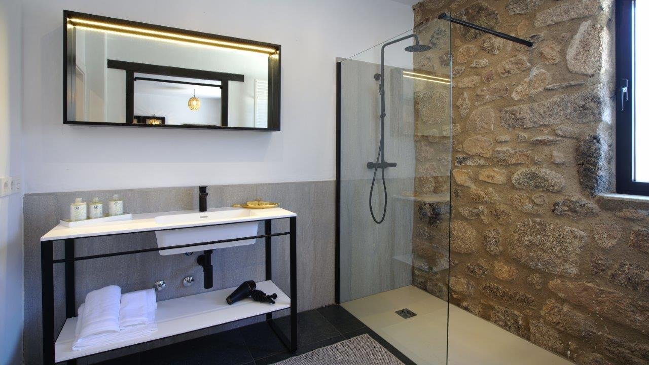 Modern bathroom with a black metal-framed vanity, white sink, mirror, and a glass-enclosed shower area with stone wall and a window.