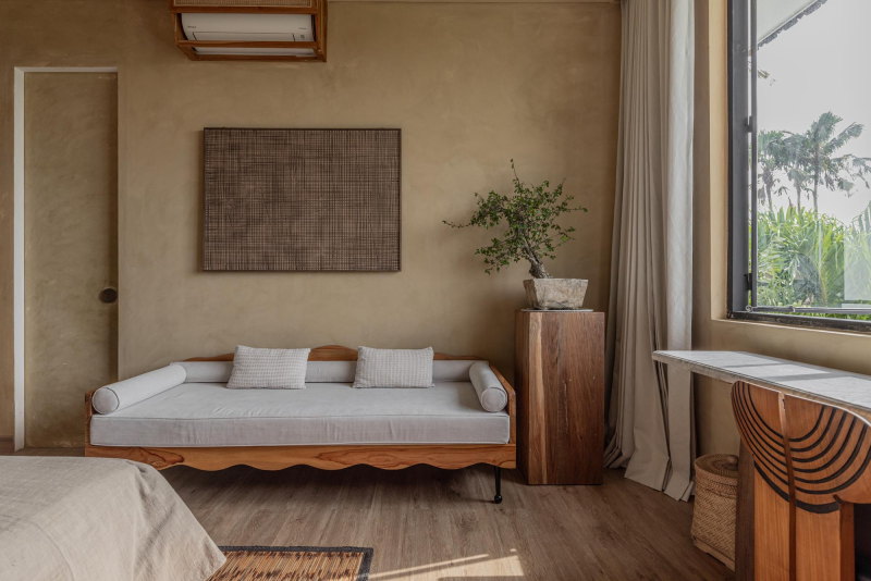 A cozy living room with a cream sofas, a large window with white curtains, a potted plant on a wooden stand, and a desk with a rounded chair. The room has beige walls and wooden flooring.