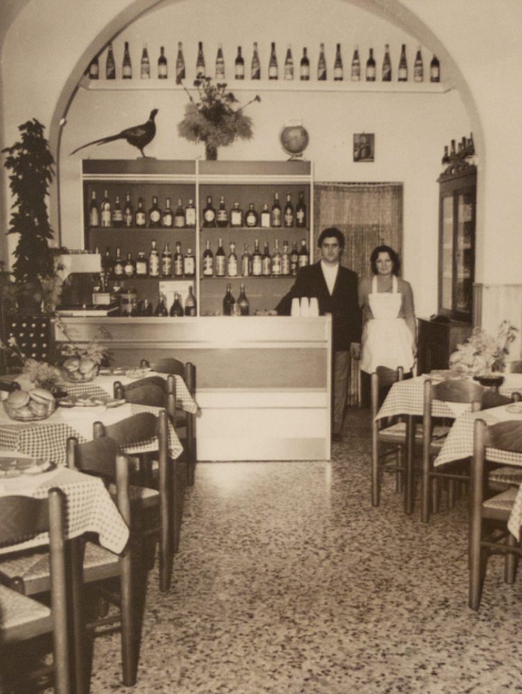 Vintage black-and-white photo of a restaurant interior with tables covered in checkered tablecloths, a bar with shelves filled with bottles, two waitstaff standing behind the bar, and decorative items including a bird sculpture, a plant, a globe, and framed pictures.