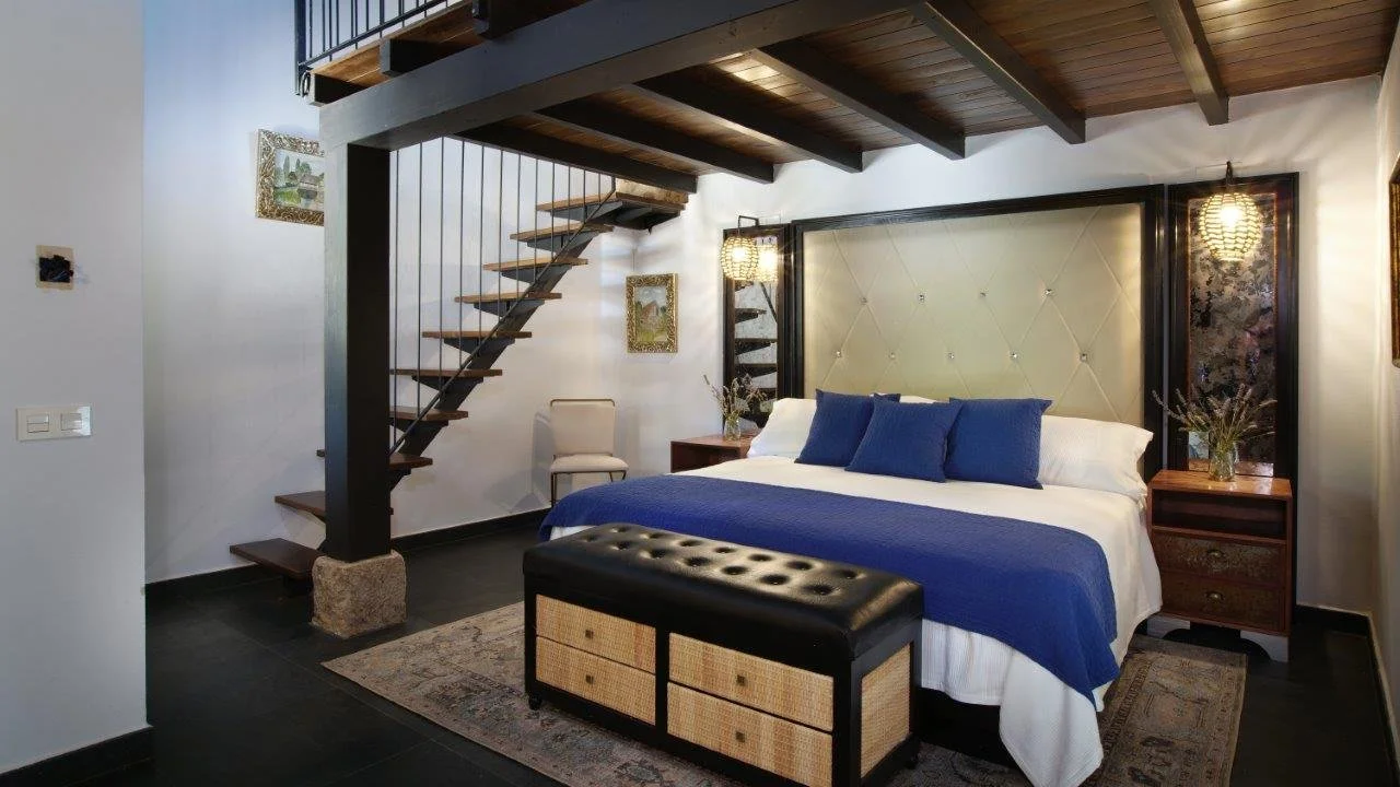 A bedroom with a large bed, blue throw blanket, and matching blue pillows. Two wooden nightstands with lamps and decorative vases, an upholstered headboard, a black leather and wicker storage bench, a patterned rug, and a wooden staircase in the background leading to a loft area.