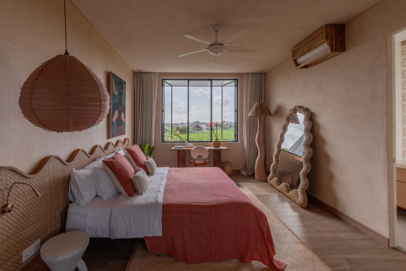 A bedroom with a large window showing green fields outside, a bed with white sheets and pinkish-red bedding, decorative pillows, a rattan headboard, a hanging paper lantern, a standing mirror, a floor lamp, a small round side table, and an air conditioning unit on the wall.