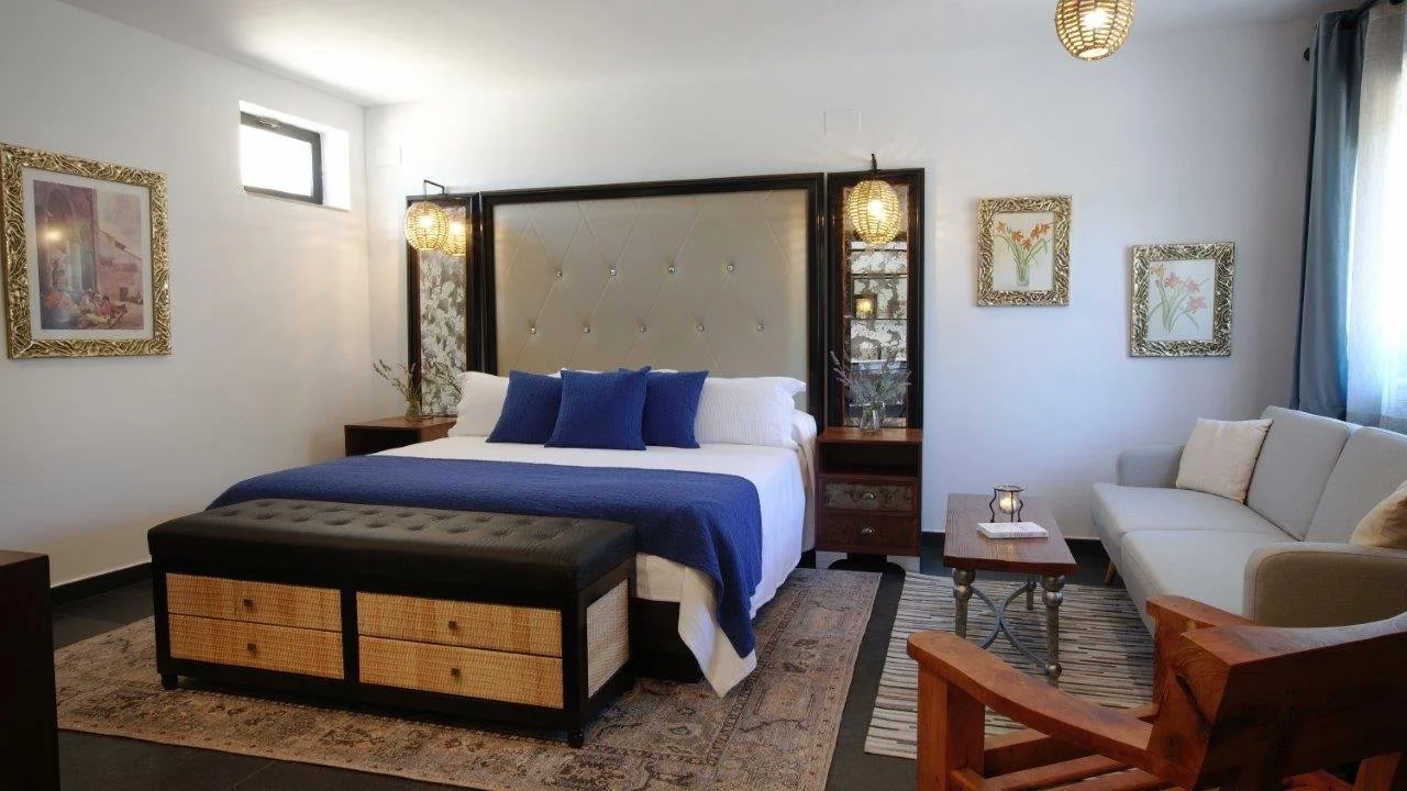 hotel room with a large bed, navy blue bedding, pillows, a bench at the foot of the bed, a white sofa, a wooden coffee table, artwork on the walls, and curtains on a window.