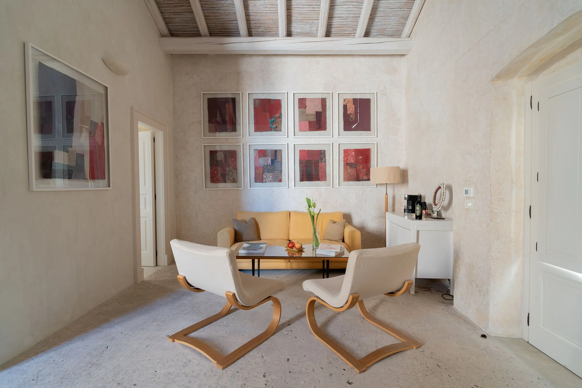 Living room with a yellow sofa, two beige armchairs, and a coffee table with books, a vase with green plant, and fruits. Wall art with nine framed red abstract pieces. Side white cabinet with a lamp, coffee maker, and decorative items. Light-colored walls and ceiling with wooden beams.