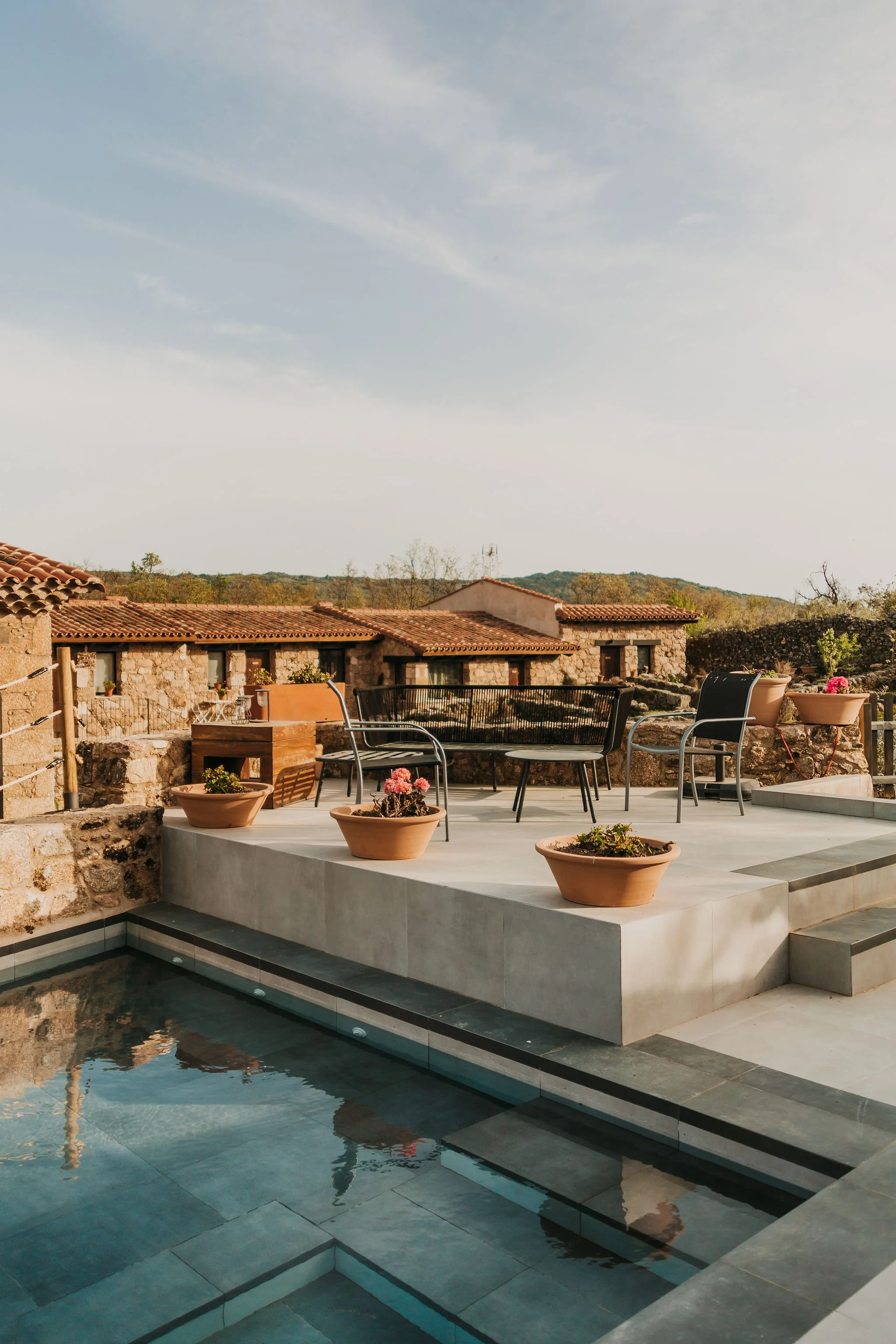 Outdoor terrace with potted plants, black outdoor furniture, and a swimming pool in a rustic setting with stone buildings and a cloudy sky.