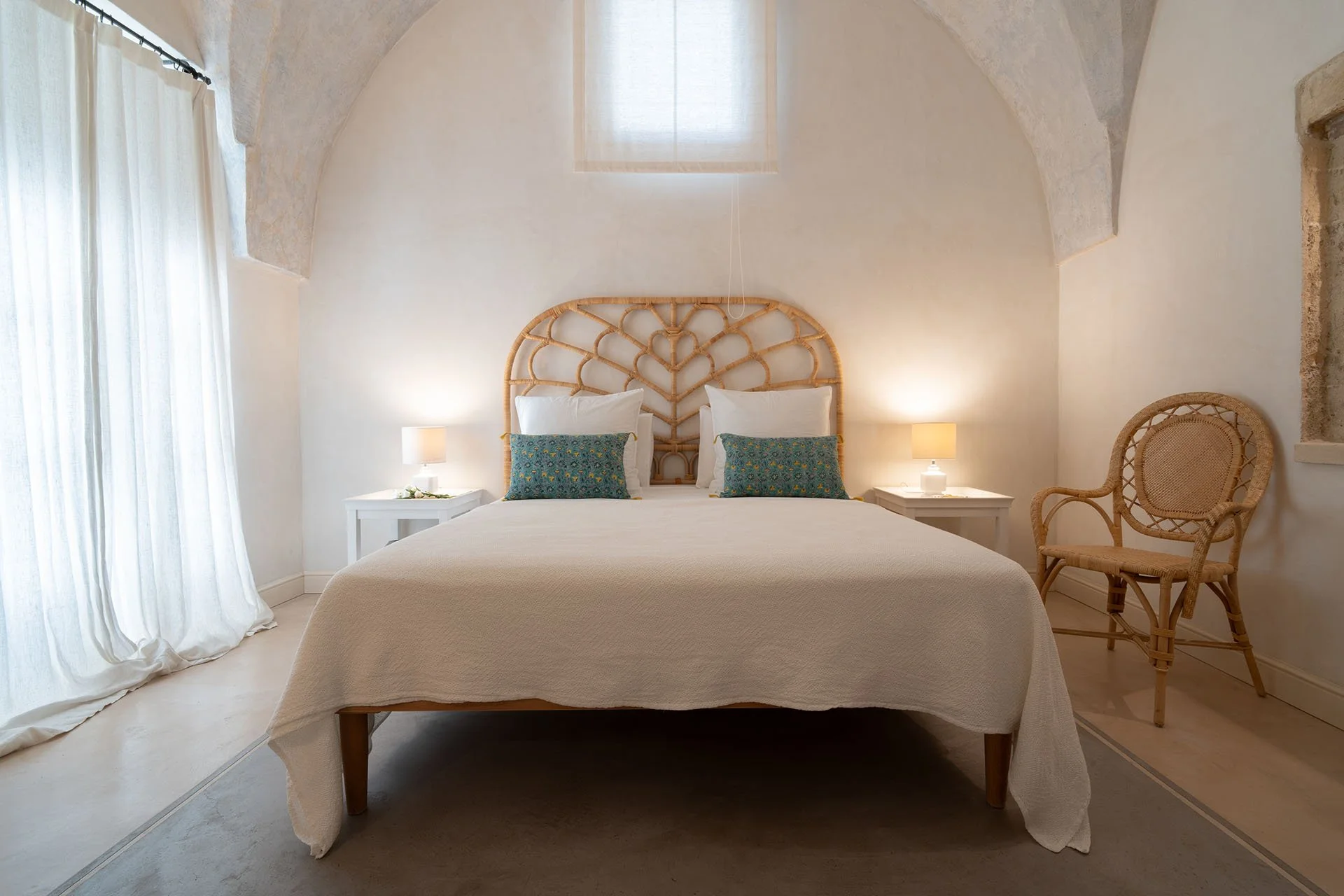 A minimalist bedroom with a king-sized bed, bamboo headboard, two nightstands with lamps, a wicker chair, white curtains, and a small window in a room with white walls and high arched ceiling.