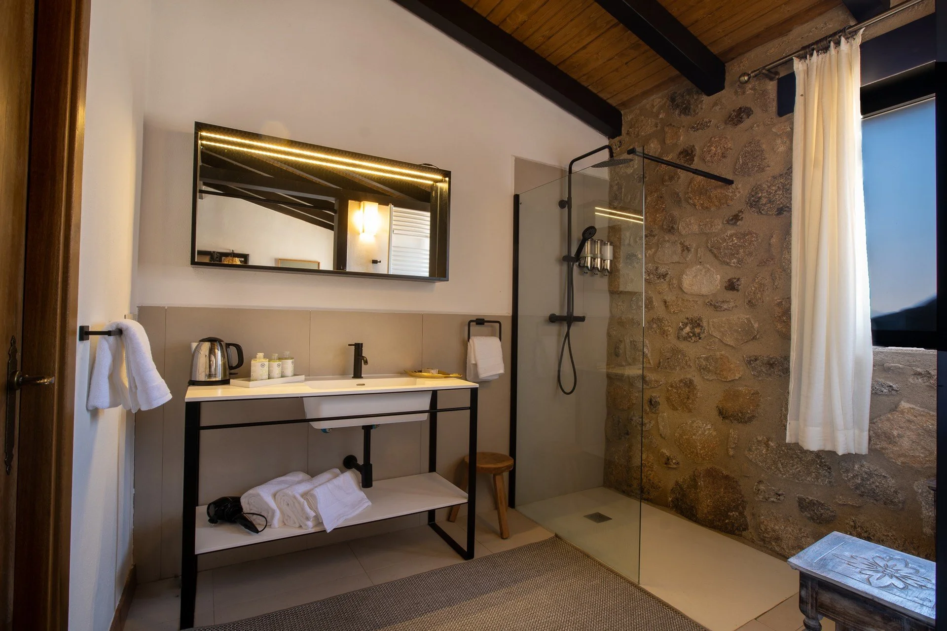 Modern bathroom featuring a glass-enclosed walk-in shower with stone wall, white sink with black fixtures, large mirror, white curtains, and natural lighting.