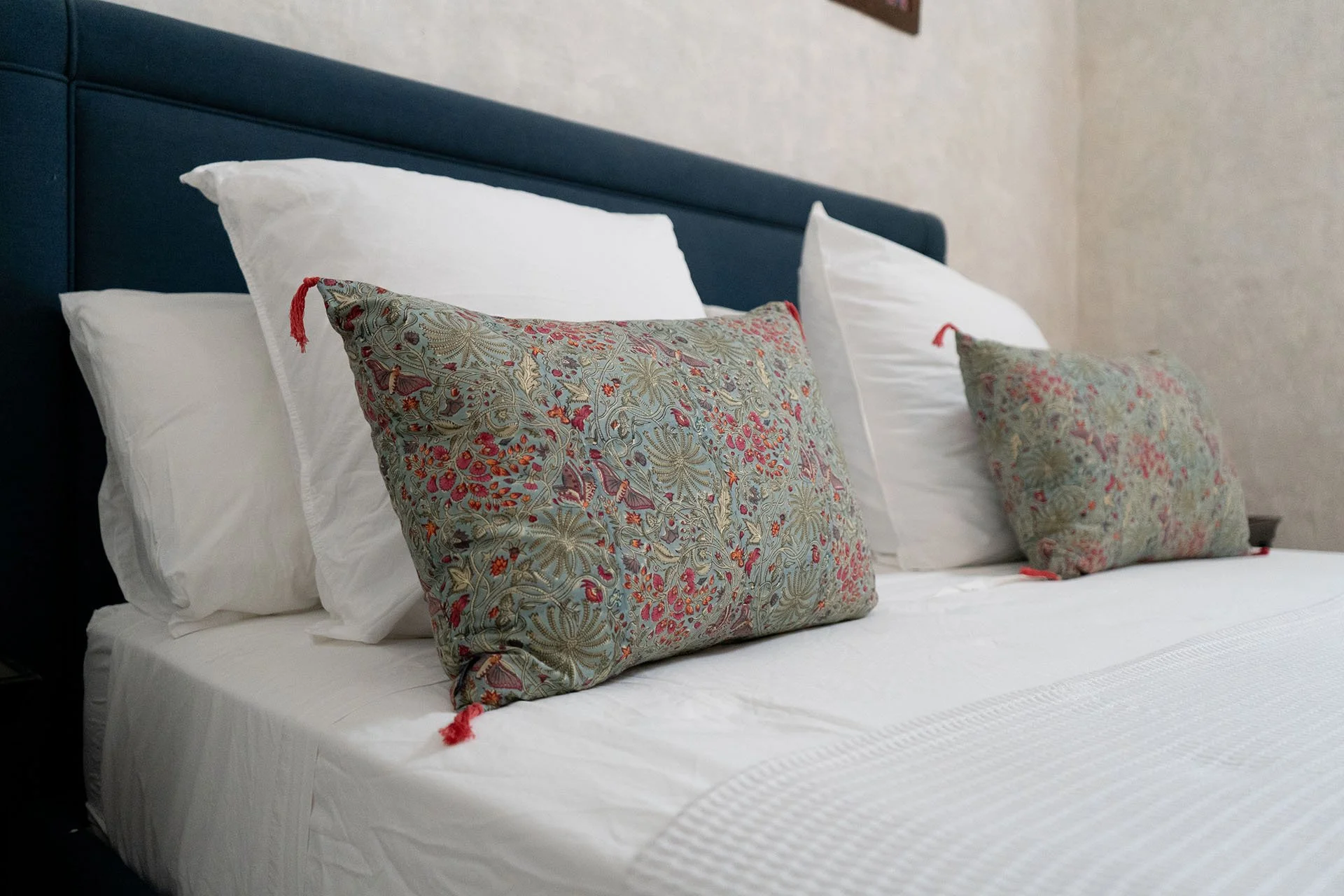 A bed with white sheets and pillows, and two decorative pillows with floral patterns in front of white pillows, against a blue headboard in a hotel room.