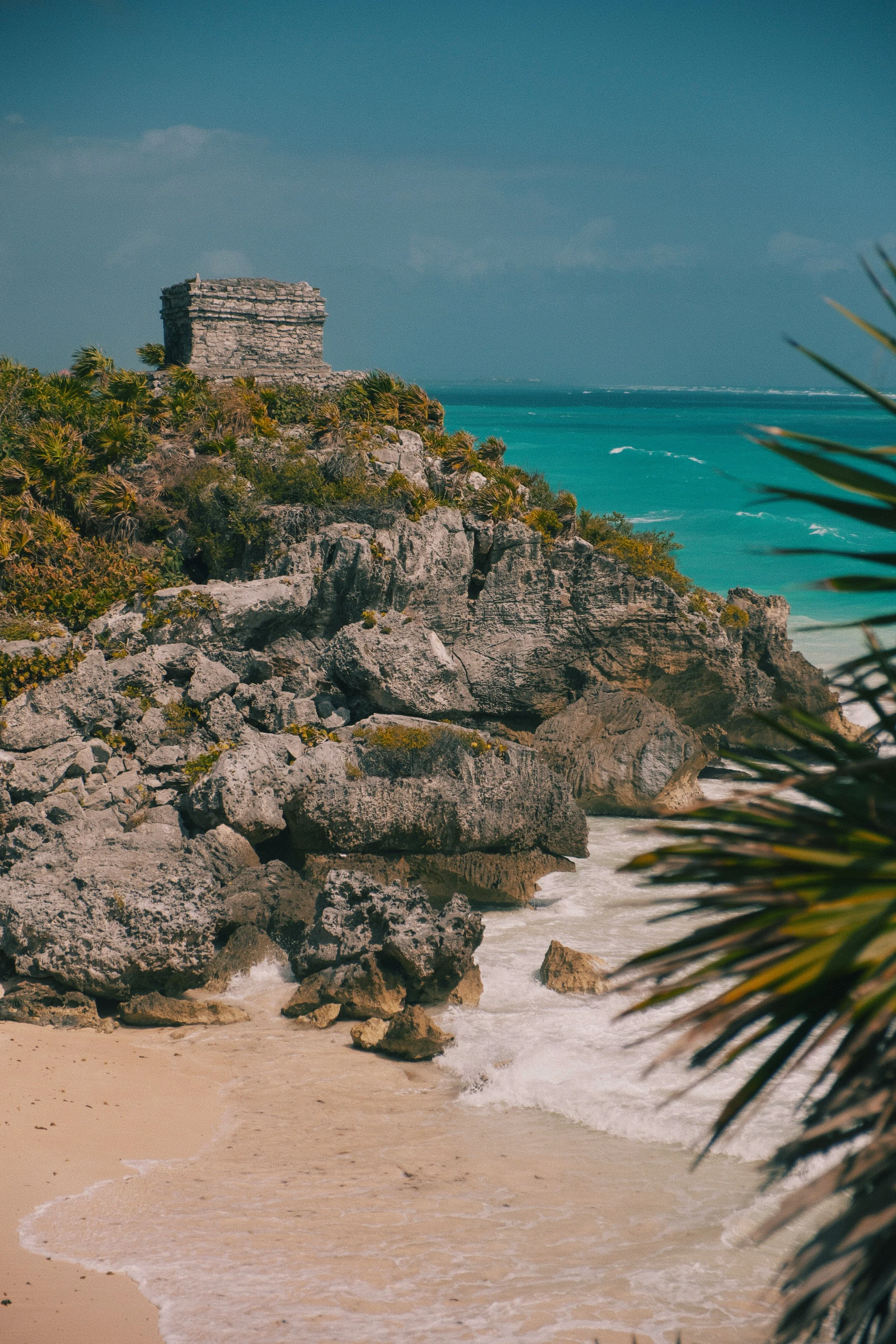 A tropical beach with sandy shore, turquoise waters, rocky cliffside, lush vegetation, and a small ancient stone structure on the cliff top.