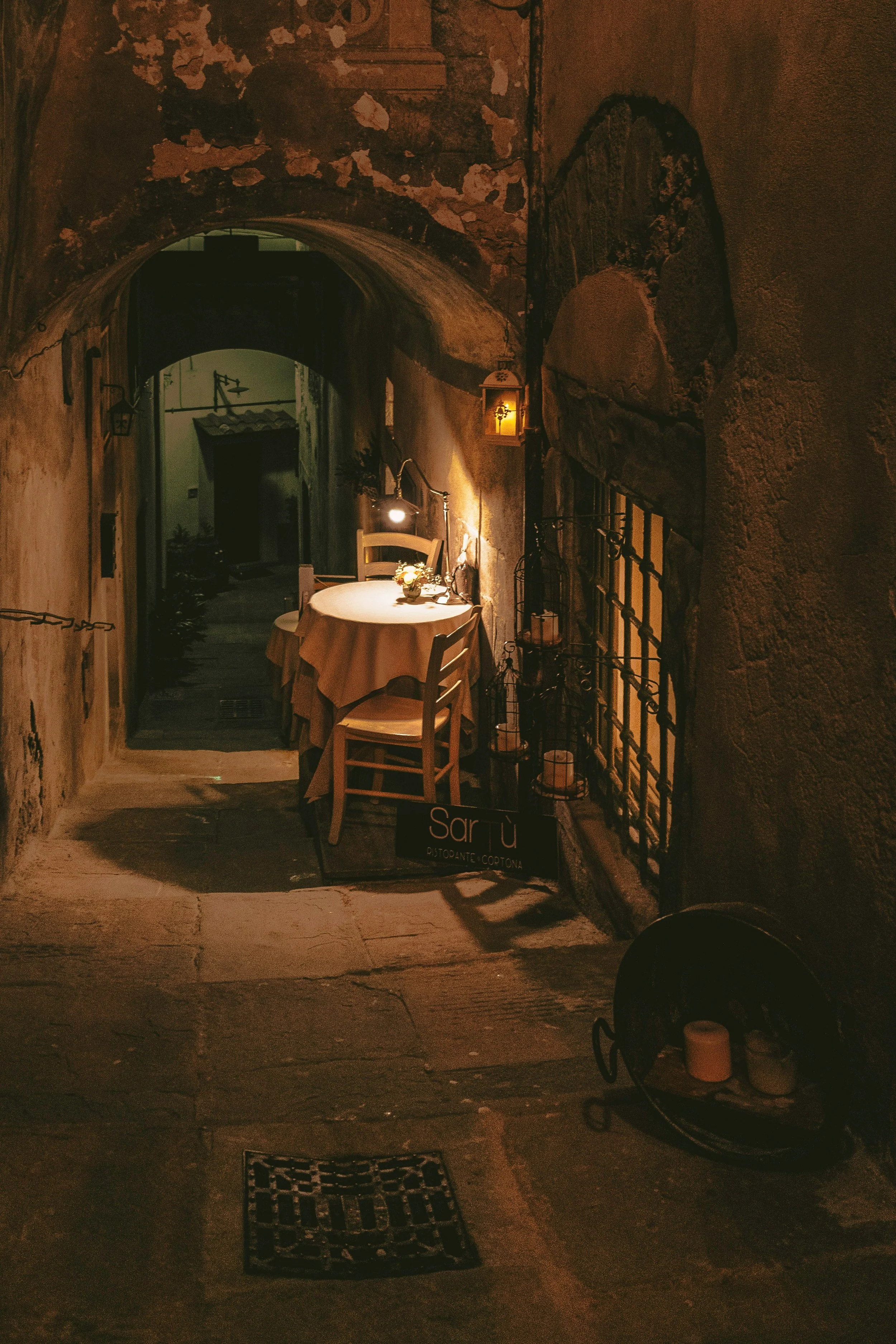 Cozy dimly lit alley with a small round table, chairs, and candles, leading to a rustic building entrance.