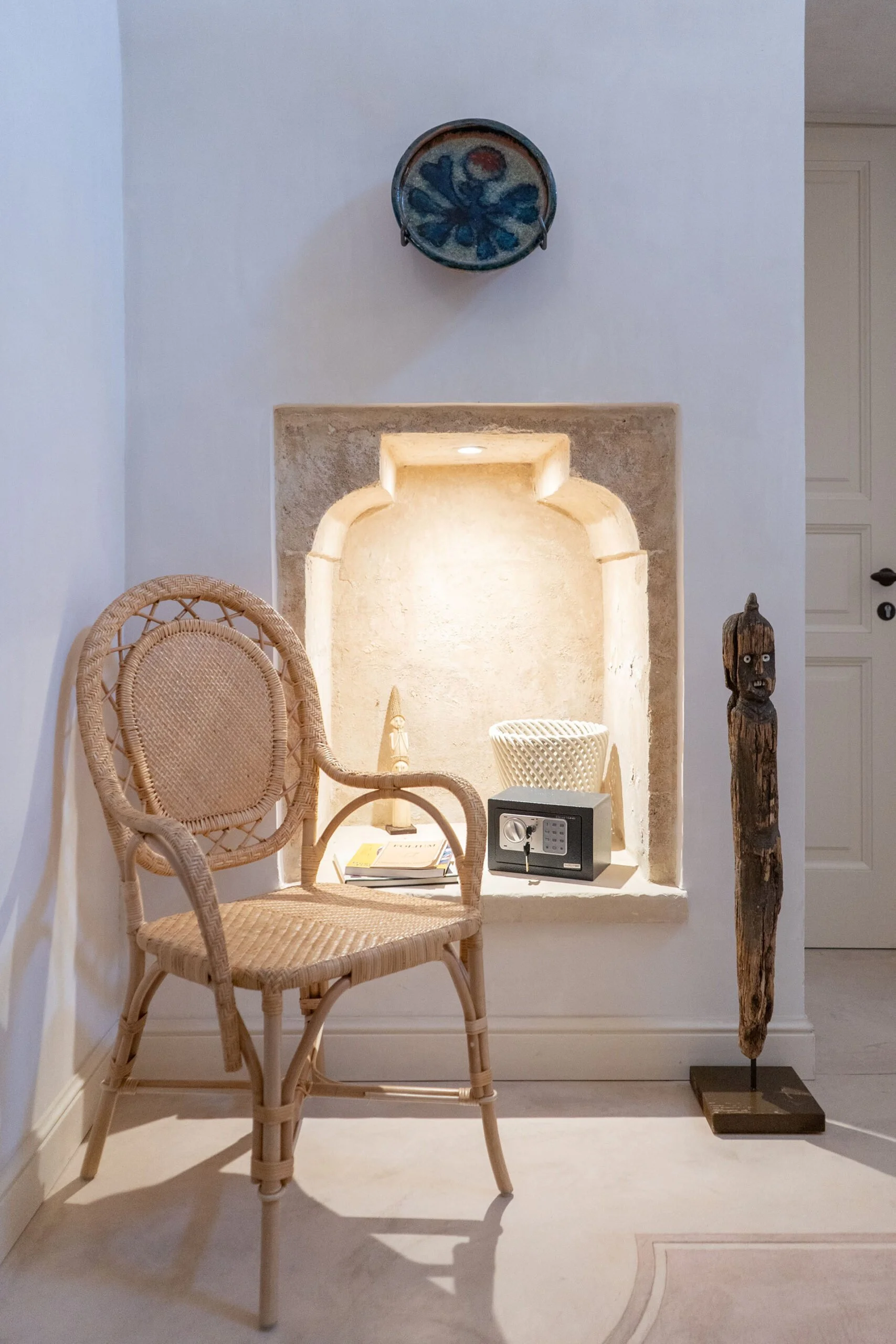 Interior corner with a rattan chair, a small safe, a decorative wooden sculpture, and display items in a niche with warm lighting, decorated with a ceramic wall hanging above.