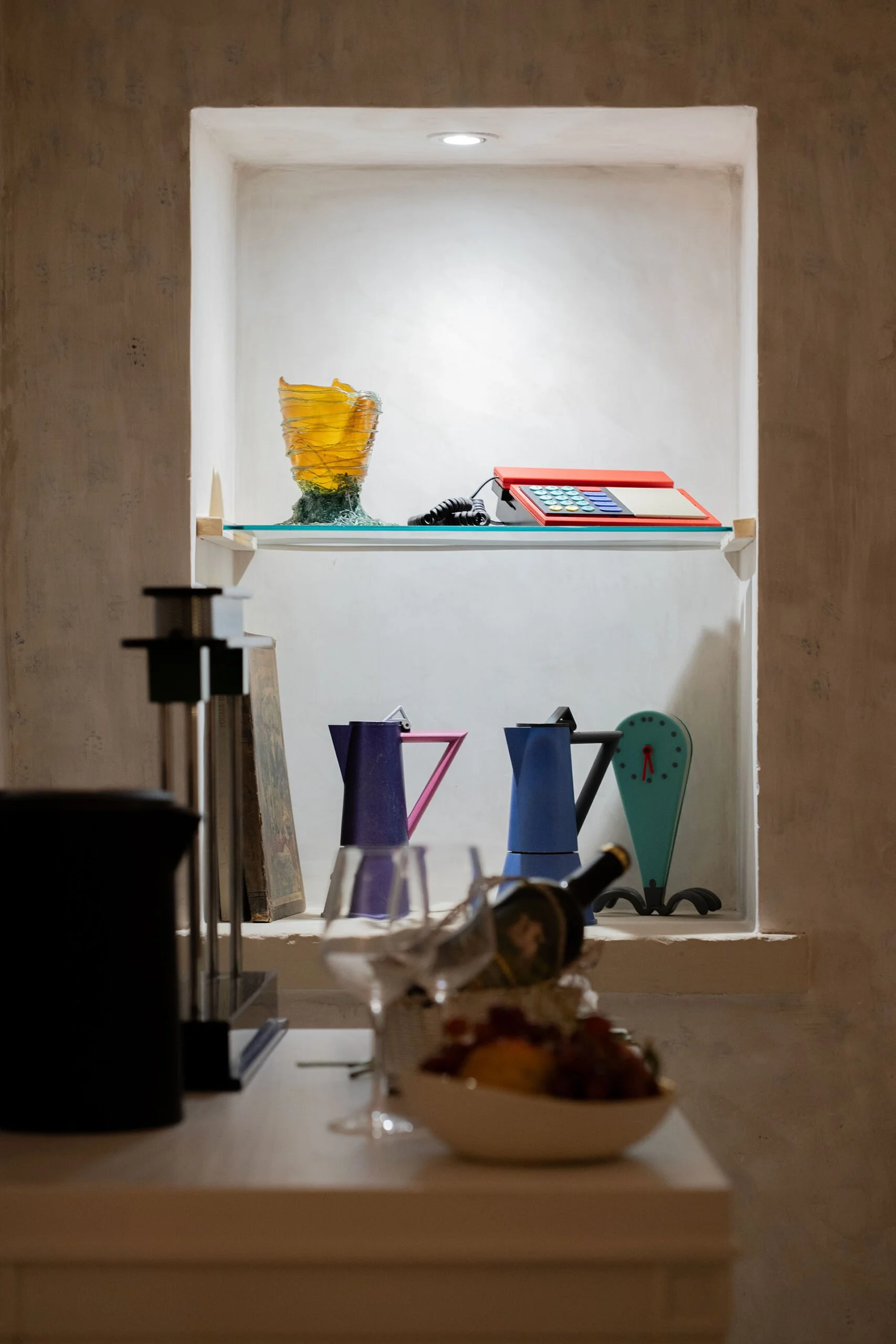 A white shelf with decorative items, including a yellow glass vase, a vintage red telephone, and colorful kitchen timers, with a bowl of food and a bottle in the foreground.