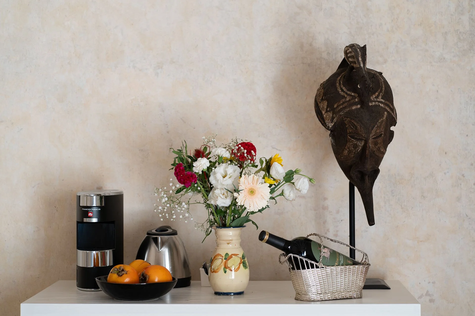 A tabletop with a fruit bowl, a coffee machine, a kettle, a vase of flowers, a wine bottle in a basket, and a wooden African tribal mask on a stand, against a beige wall.