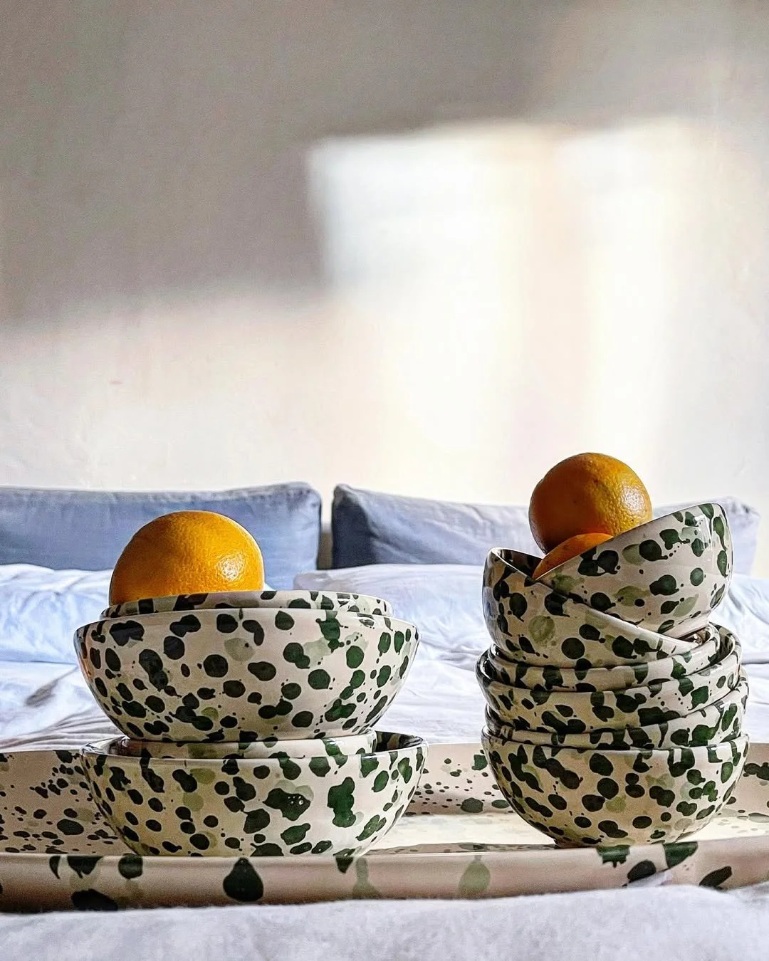 Two sets of scalloped ceramic bowls with black and green splatter pattern, stacked with oranges on top, placed on a matching splatter tray in front of a bed with white and blue pillows, against a plain wall.
