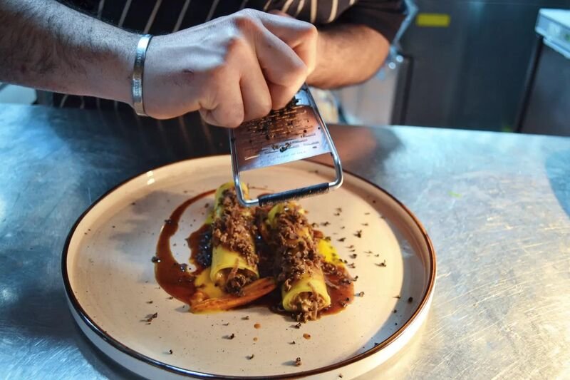 Person grating chocolate over a plated dish with stuffed rolls topped with sauce and herbs.