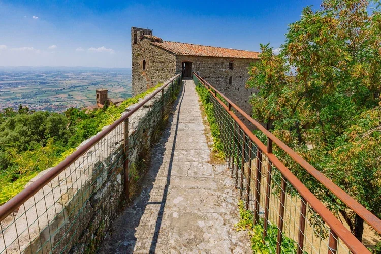 Stone walkway with rusted metal railings leading to an old stone building on a hilltop, overlooking a green landscape and distant cityscape under a blue sky.