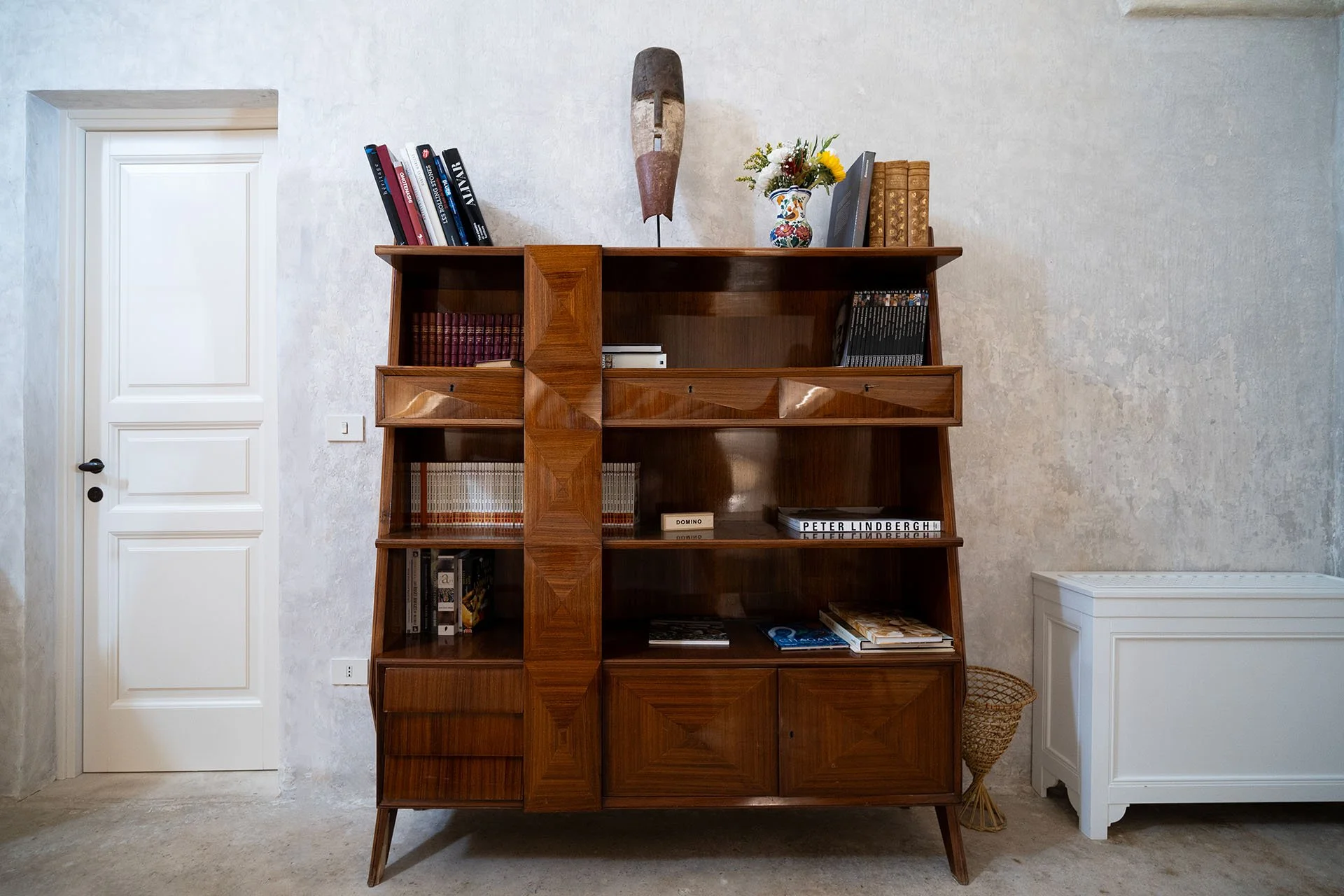 Mid-century modern wooden bookshelf with various books, a vase with flowers, and decorative items, against a textured white wall.