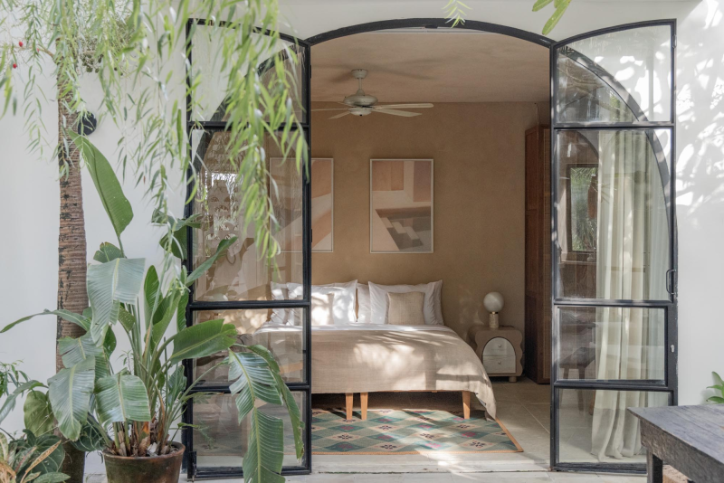 View of a cozy bedroom seen through black-framed glass doors, with a bed, artwork, and greenery outside.