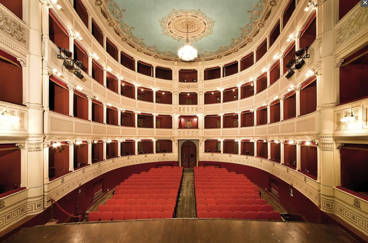 View of an elegant, empty theater with red velvet seats and ornate white and gold balconies, seen from the stage.