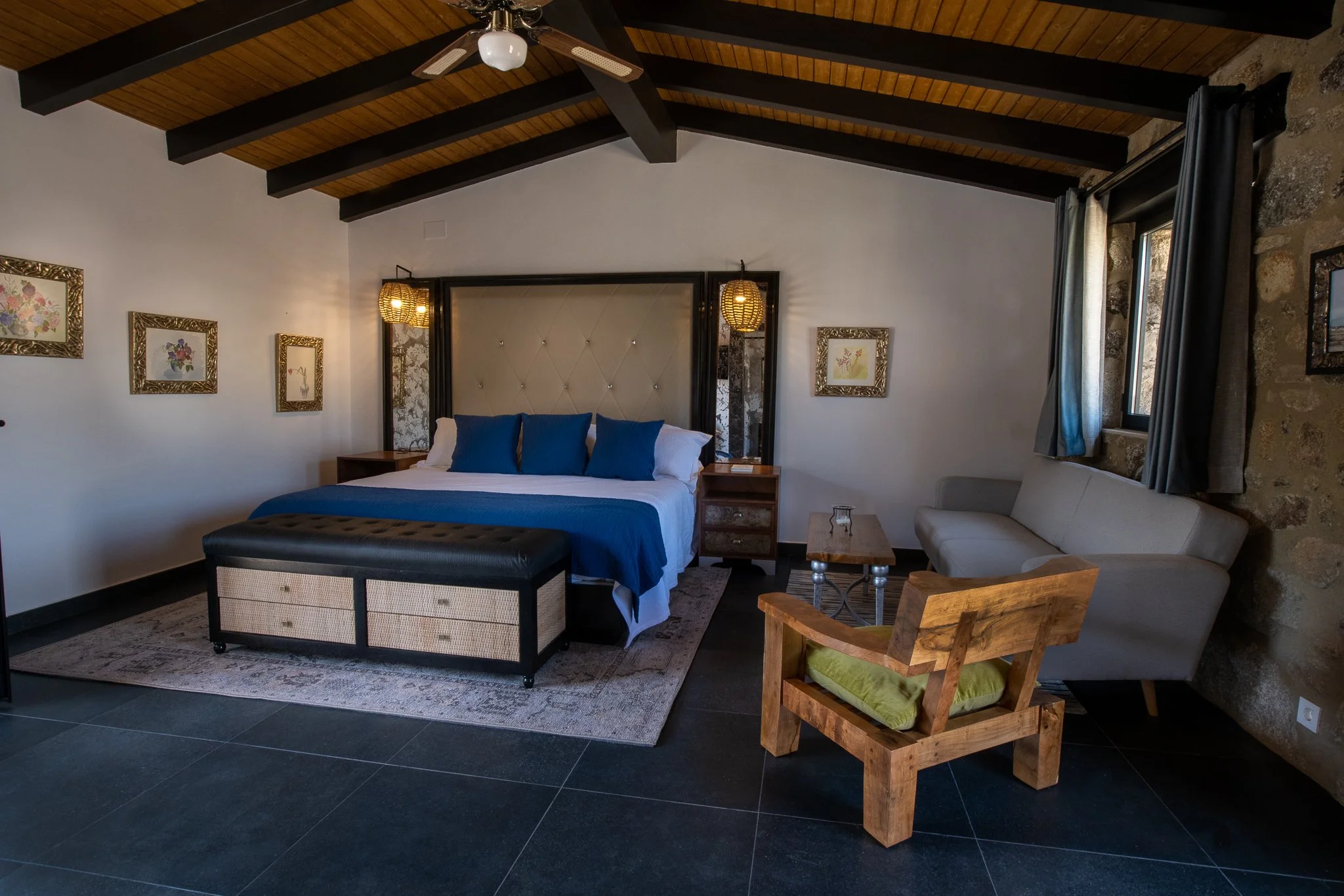 A bedroom with a high wooden ceiling, a bed with blue pillows and a beige tufted headboard, a black and beige storage bench at the foot of the bed, a gray sofa, a wooden armchair with a green cushion, a small wooden coffee table, and framed floral artwork on the walls.