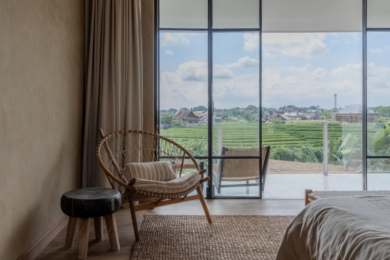 A cozy bedroom with large floor-to-ceiling glass windows overlooking green fields and a distant town, a wicker chair with a cushion, a small black ottoman, and a bed partially visible on the right.