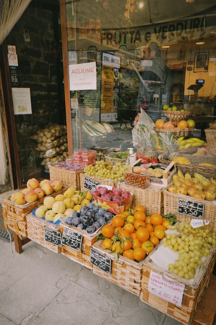 Display of various fruits at a market stand, including peaches, grapes, oranges, and bananas, with price tags and a storefront window behind.