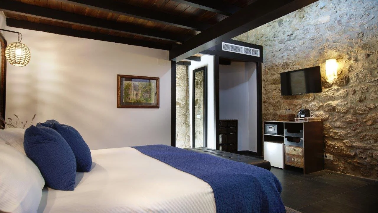 Room with stone and white walls, a bed with white linens and blue pillows, a wall-mounted TV, and a small cabinet with a safe on top.