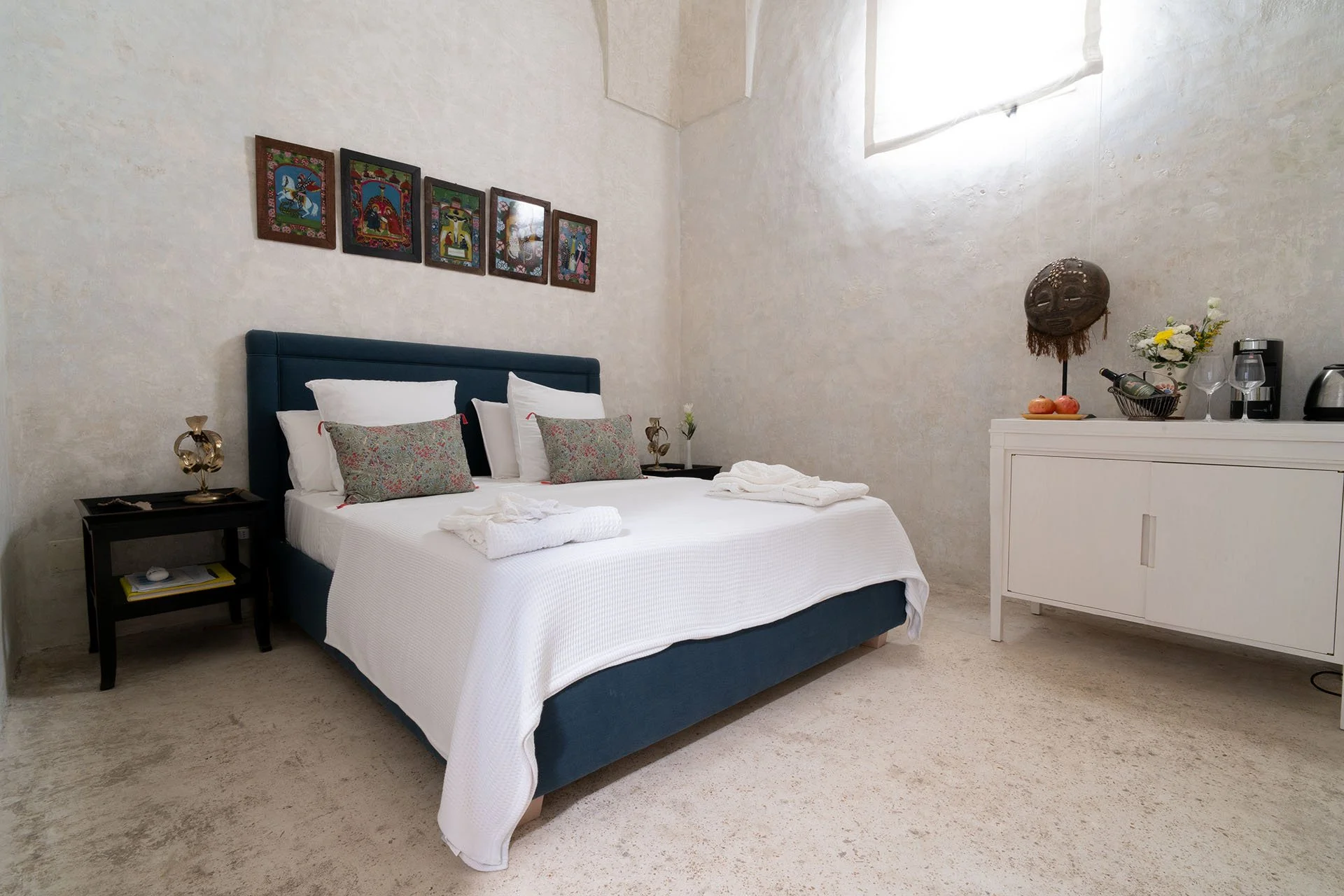 A bedroom with a blue upholstered bed, white bedding, and decorative pillows. There are three small beds on each side of the bed with matching lamps. A collection of five colorful framed artworks hangs above the bed. To the right, there is a white cabinet with a flower vase, fruit, coffee machine, and other items on top. The walls are textured and the room is illuminated by a window near the ceiling.