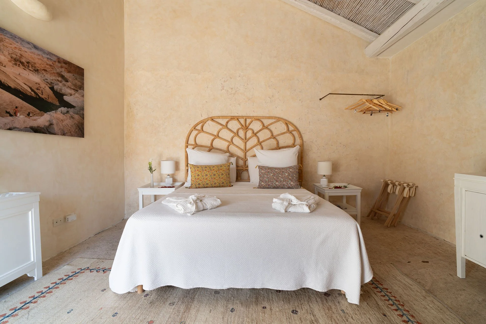 A cozy bedroom with a large bed featuring a woven headboard, white bedding, and decorative pillows. There are two white nightstands with lamps on either side of the bed, and a painting on the wall to the left. An open closet with hanging wooden hangers is on the right, and a small wooden bench is in the corner. The room has warm, beige walls and a light-colored carpet.