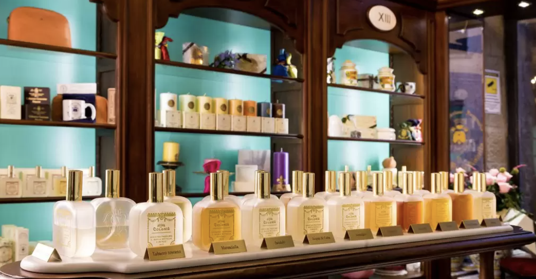 Display of various perfume bottles with gold caps on a white surface, set against a teal wall with shelves holding candles and other items.