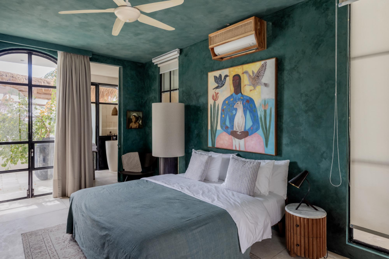 A bedroom with dark teal walls, a white bed with gray pillows, a large colorful painting featuring a figure with birds and animals, a white ceiling fan, and a window with beige curtains revealing outdoor plants.
