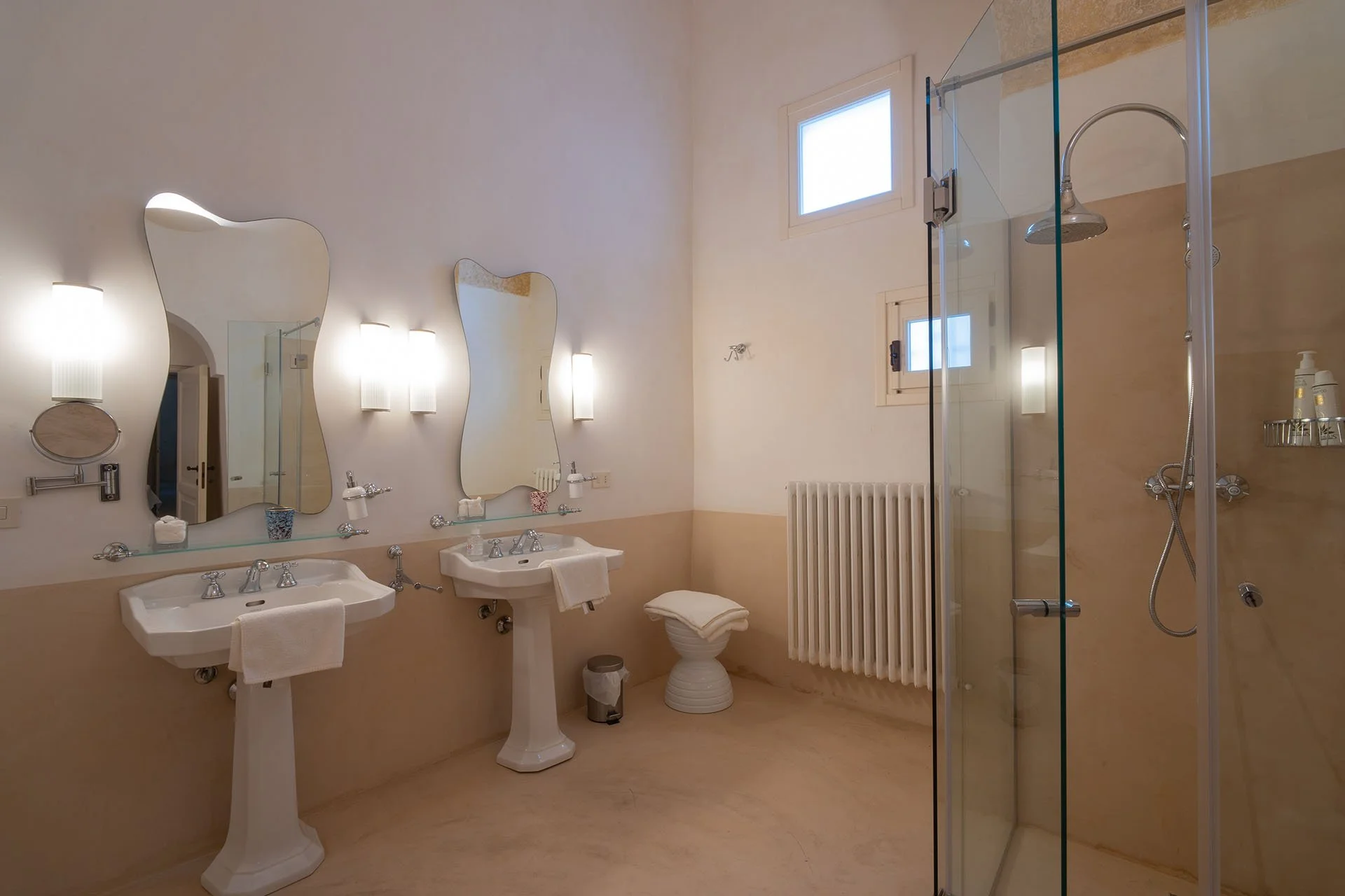A spacious bathroom with two pedestal sinks, irregularly shaped mirrors, frosted wall sconces, a small window, a radiator, and a glass-enclosed shower.