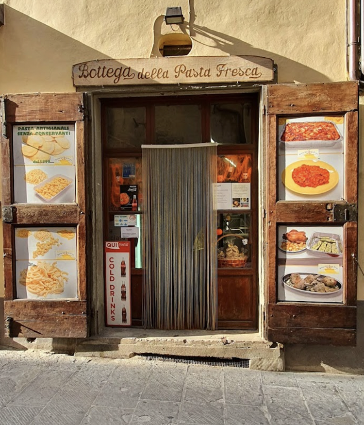 Small Italian pasta shop with a wooden sign reading "Bottega della Pasta Fresca" on top. The storefront has pictures of various pasta dishes and food items displayed on the sides.