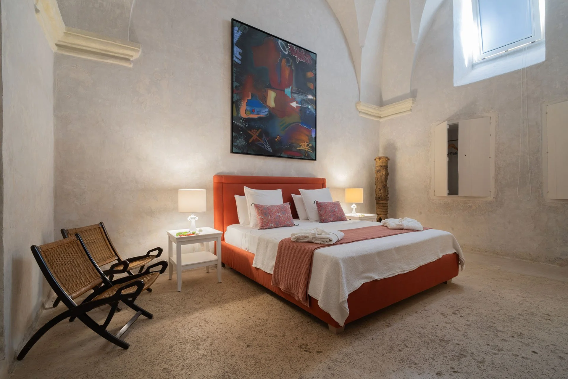 Hotel bedroom with an orange upholstered bed, white bedding, pink throw, and decorative pillows, flanked by two lamps on white nightstands, with chairs and a painting on the wall.