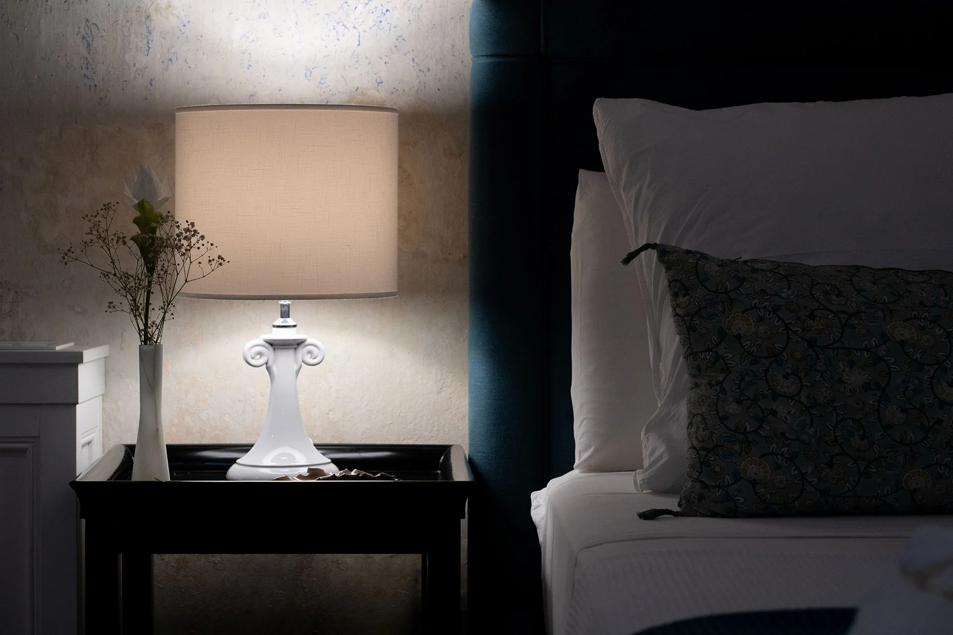 A bedside table with a white table lamp, a white vase with flowers, next to a bed with white sheets and dark patterned pillows.