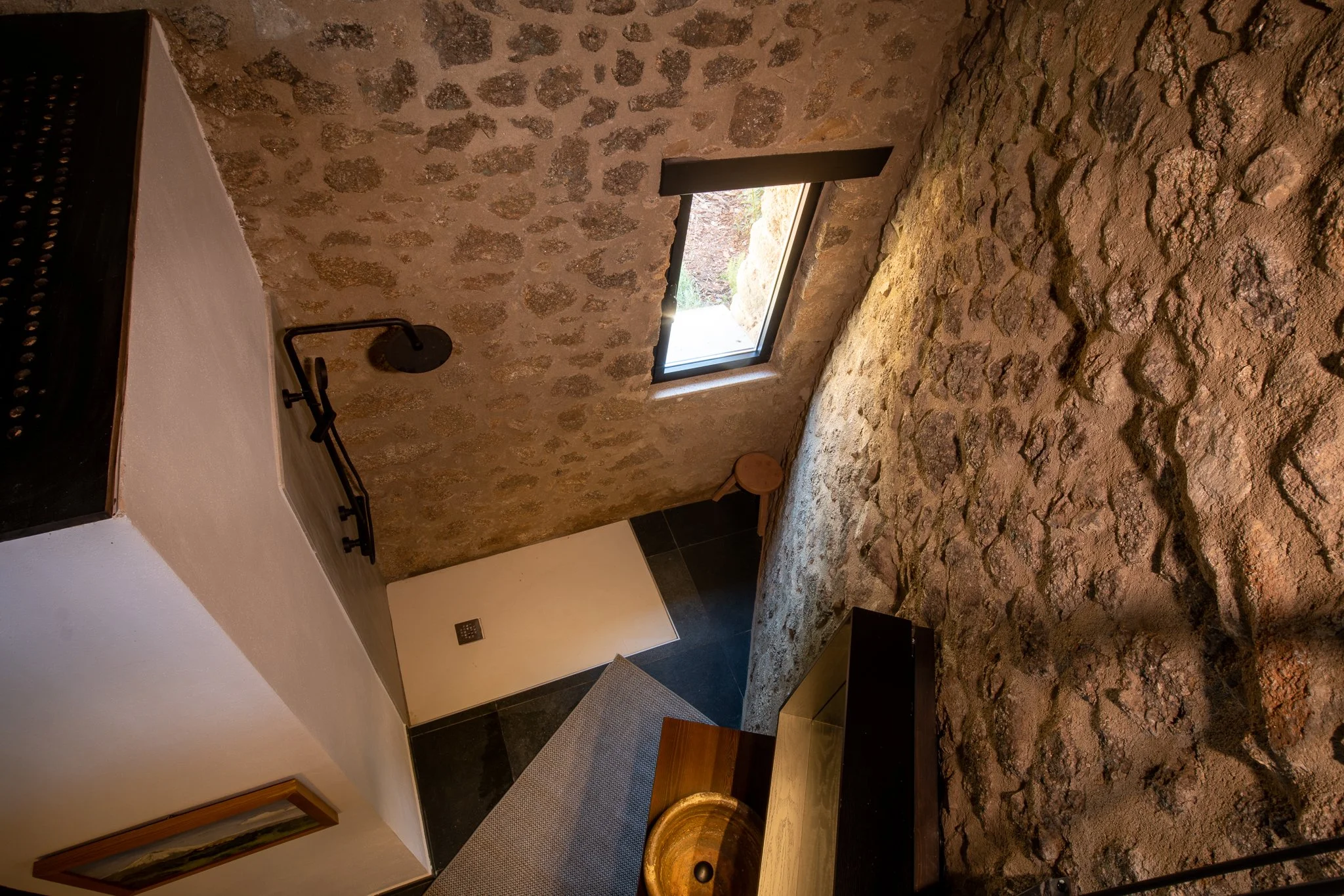 Interior of a room with rustic stone walls, a window allowing natural light, a black showerhead on the wall, part of a bed or furniture with a black frame, and a bathroom with a sink and a mirror.