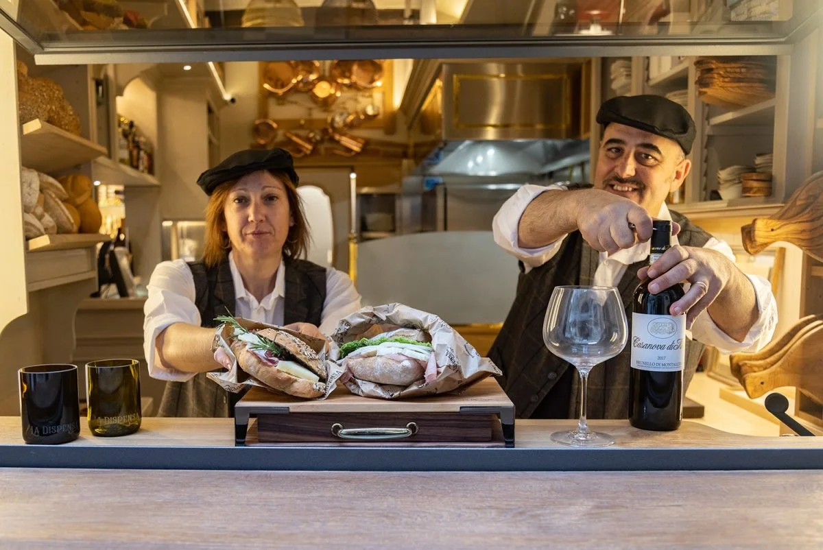 Two cafe workers, a woman and a man, behind a counter with sandwiches, wine, and glasses. The woman holds a sandwich, while the man is opening a bottle of wine. Both are wearing black berets, and the background shows a cozy kitchen with shelves and hanging copper pots.