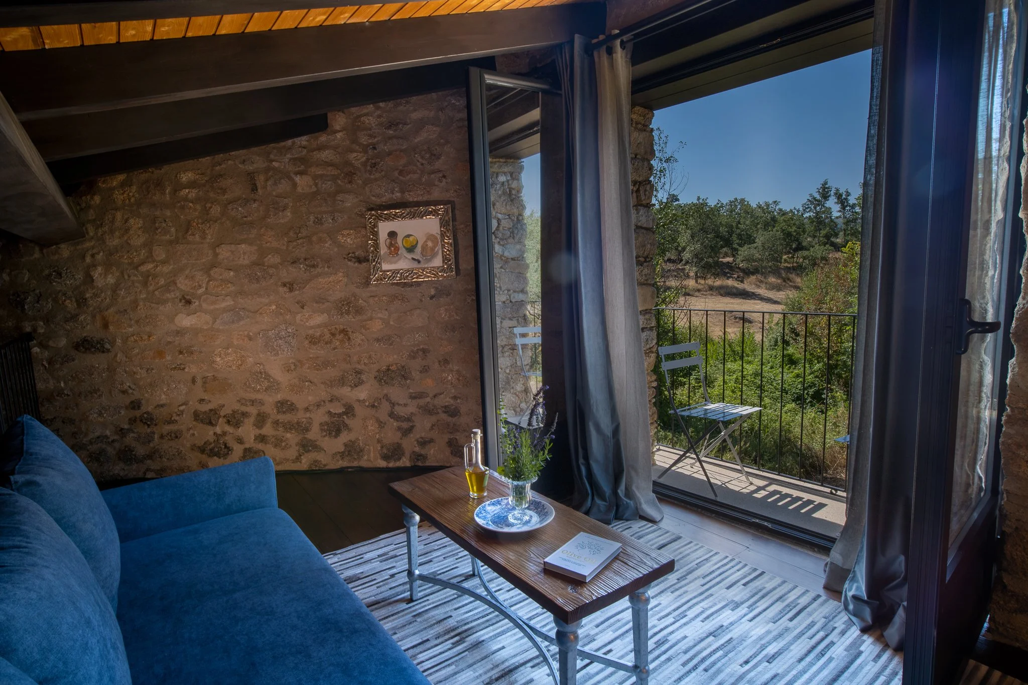 Cozy living room with stone walls, blue sofa, wooden coffee table with a book, glass bottle, and potted plant, open balcony with chairs, overlooking greenery and trees