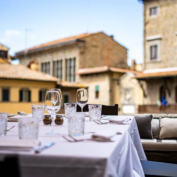 An outdoor dining table set with wine glasses, water glasses, and napkins, with rustic buildings in the background.