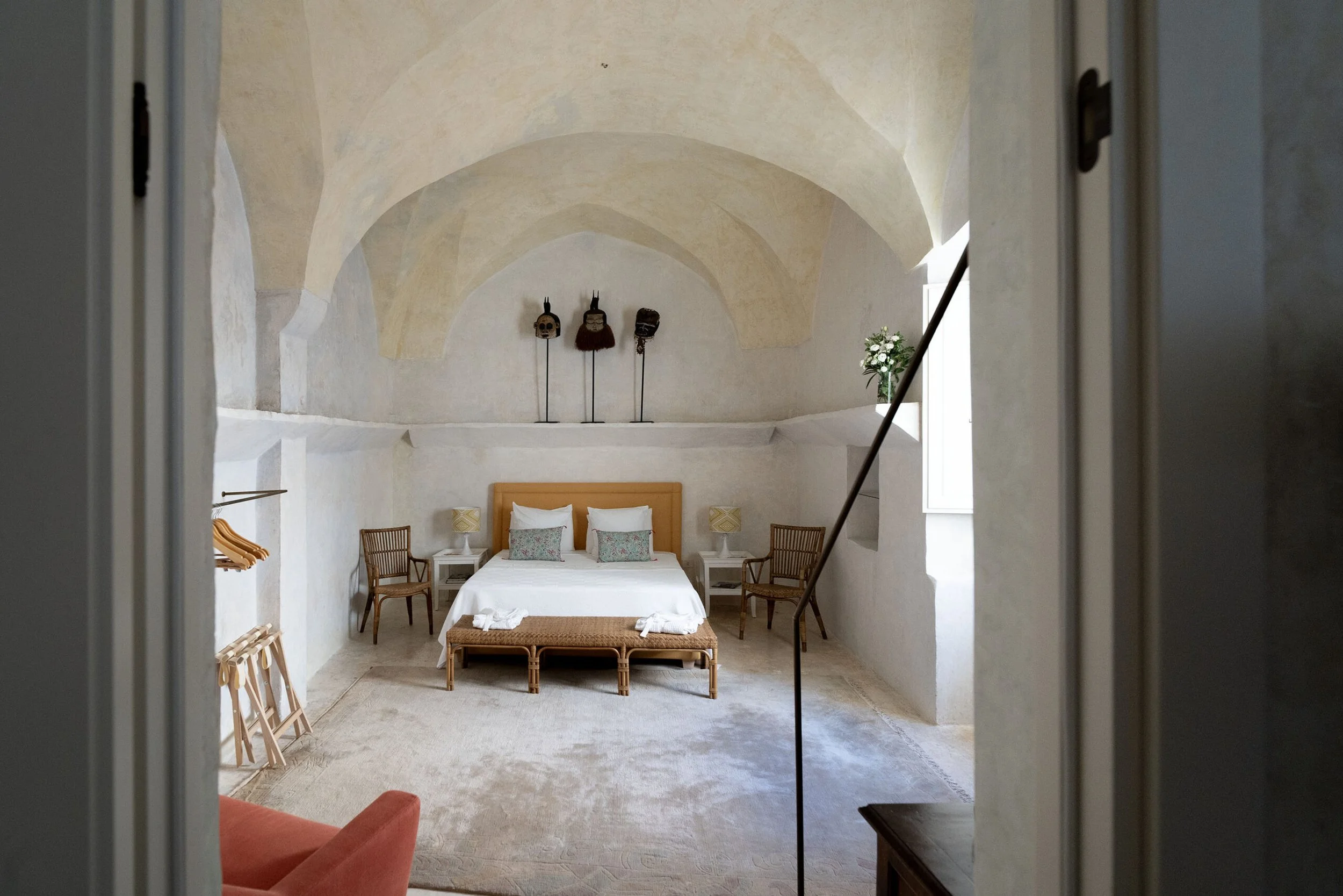 A bedroom with a vaulted ceiling, white walls, a bed with a tan headboard, white bedding, and decorative pillows. There are two nightstands with lamps, two empty chairs, a small bench at the foot of the bed, a window with sunlight, and a plant on a ledge. Artistic masks are displayed on the wall above the bed.
