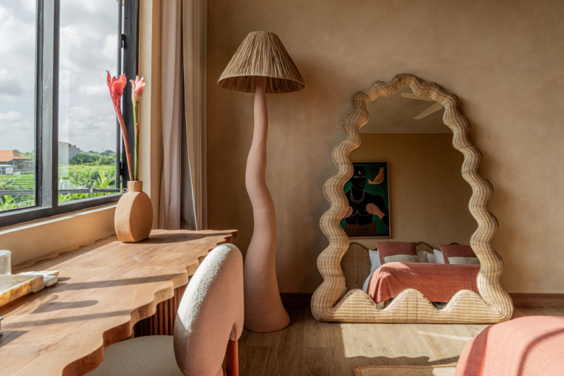 Interior of a bedroom with a unique floor lamp shaped like a human leg with a lampshade on top, a wicker mirror, a wooden desk with a vase containing pink flowers, and a window showing a scenic view outside.
